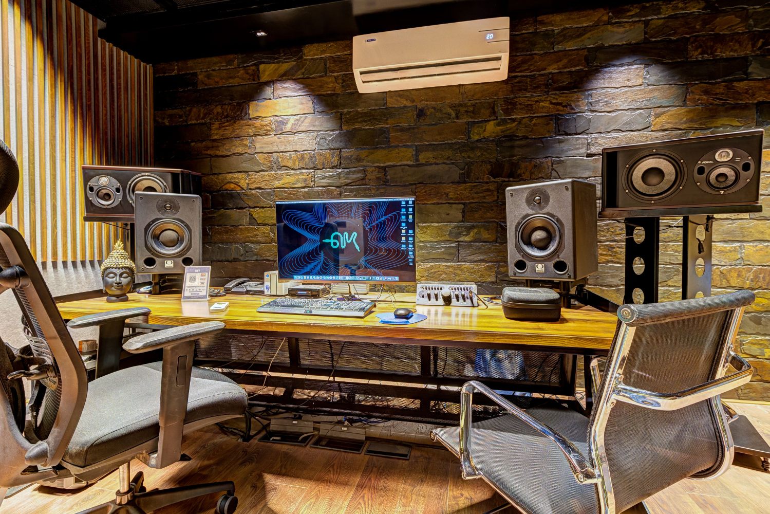 A recording studio control room with studio monitors, a mixing desk and a screen in front of a stone wall
