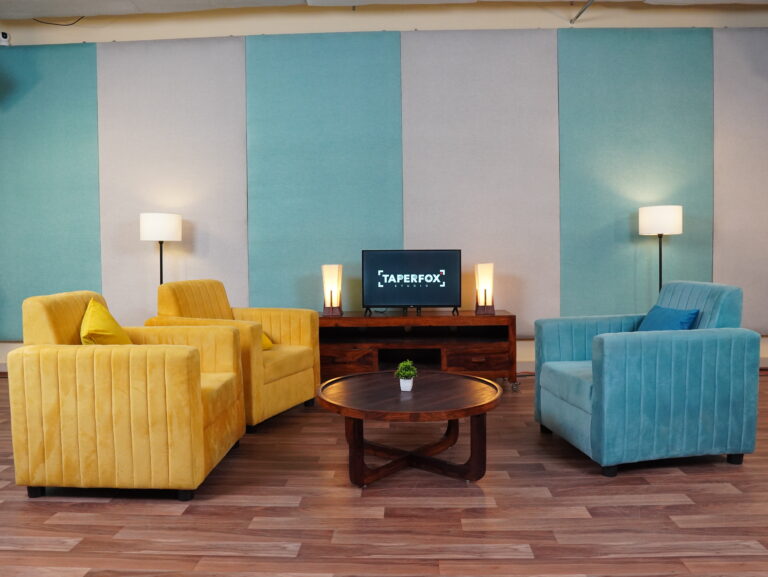 A lounge-style podcast set with yellow and blue sofas, a coffee table and acoustic panels on the wall